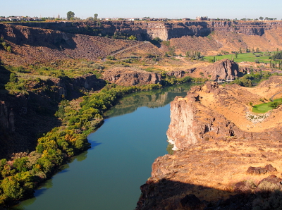 [The river curves from the right coming into view in the center of the image. To the left are many trees at the water's edge while only rock is seen to the left. A road winds down the canyon wall.]